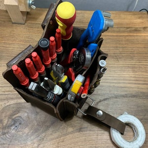 May include: A brown leather tool bag filled with various tools, including screwdrivers, pliers, wire strippers, and a yellow and red hammer. The bag has a strap and a roll of white tape is on the table next to it.