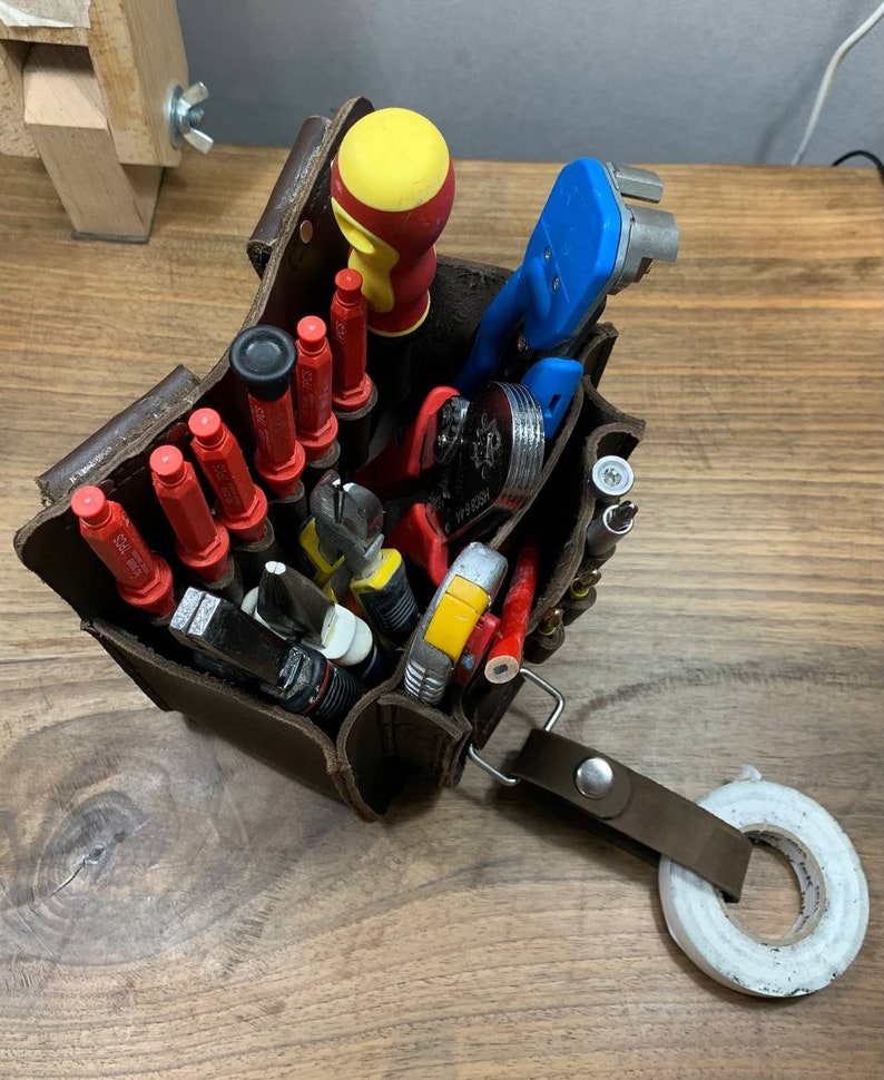 May include: A brown leather tool bag filled with various tools, including screwdrivers, pliers, wire strippers, and a yellow and red hammer. The bag has a strap and a roll of white tape is on the table next to it.