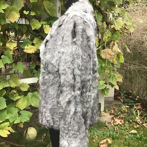May include: A gray faux fur jacket with a high collar. The jacket is on a mannequin and is in a garden setting.