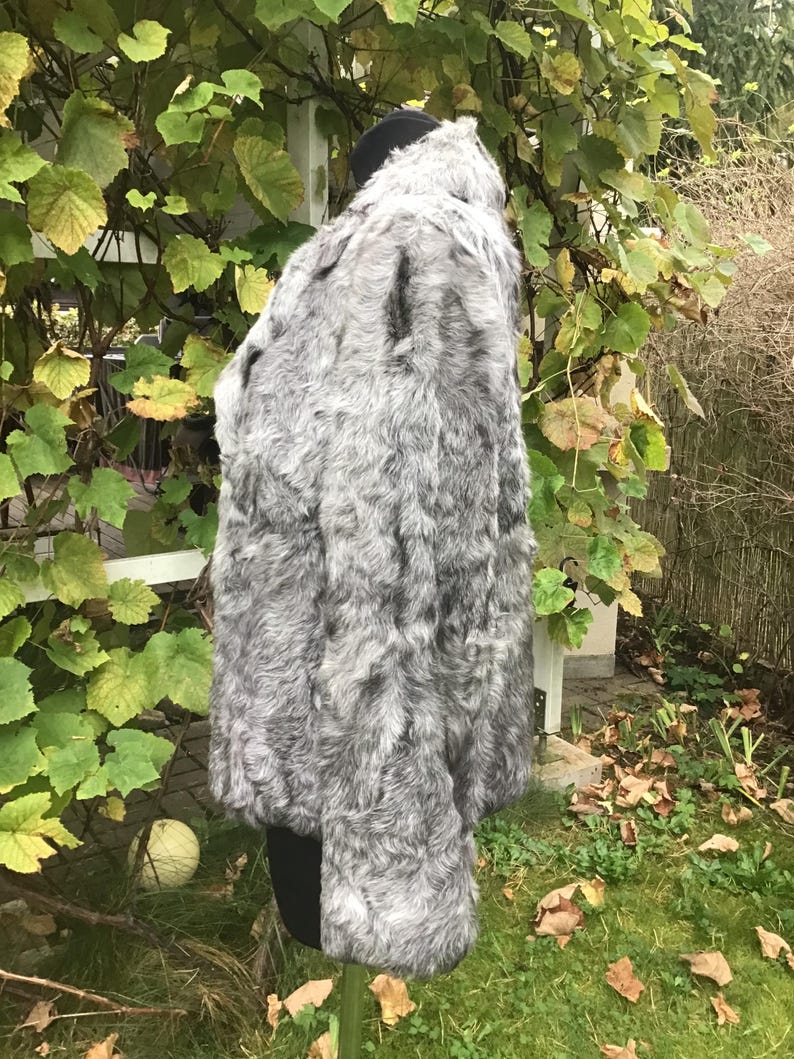 May include: A gray faux fur jacket with a high collar. The jacket is on a mannequin and is in a garden setting.