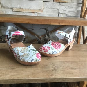 May include: A pair of white clogs with floral patterns in pink and red. The clogs have wooden soles and a strap with a buckle. The shoes are on a wooden surface.