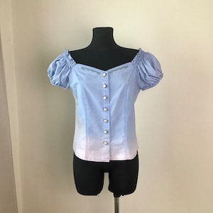 Pu&ograve; includere: Una blusa azzurra con un effetto ombr&eacute; bianco sul fondo. La blusa ha una chiusura con bottoni sul davanti, maniche corte a sbuffo e una scollatura a cuore.