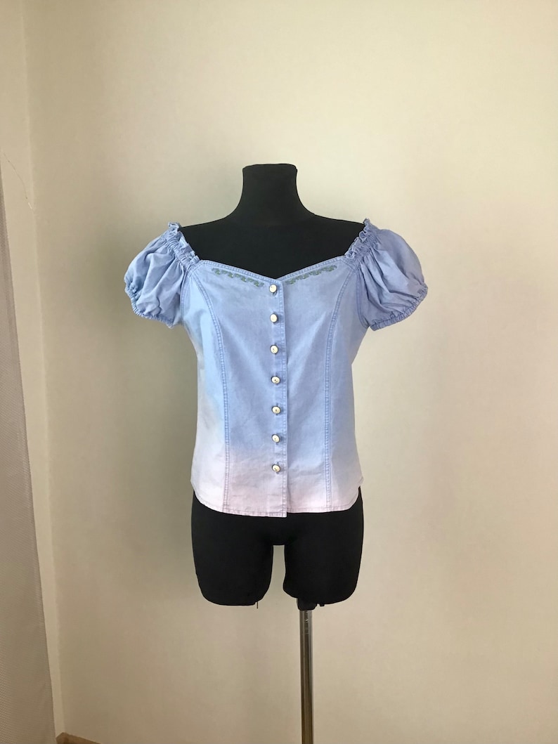 Pu&ograve; includere: Una blusa azzurra con un effetto ombr&eacute; bianco sul fondo. La blusa ha una chiusura con bottoni sul davanti, maniche corte a sbuffo e una scollatura a cuore.