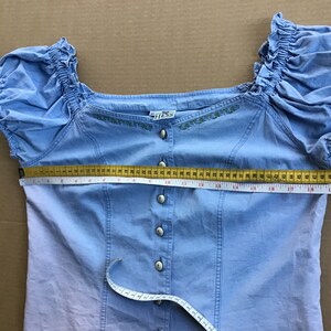 Pu&ograve; includere: Una blusa azzurra a spalle scoperte con chiusura a bottoni sul davanti e dettagli floreali ricamati. La blusa ha maniche corte a sbuffo e un corpetto aderente. La blusa viene misurata sul seno con un metro a nastro, mostrando una misura di 53,3 cm.