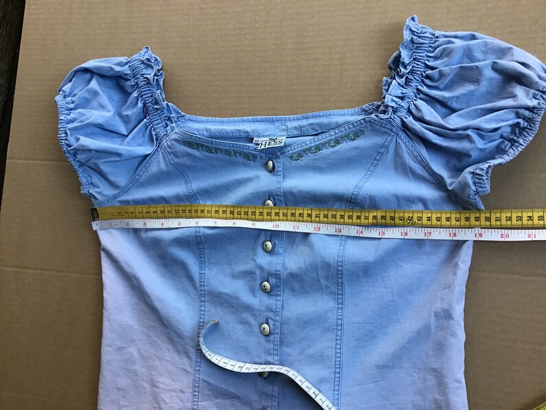 Pu&ograve; includere: Una blusa azzurra a spalle scoperte con chiusura a bottoni sul davanti e dettagli floreali ricamati. La blusa ha maniche corte a sbuffo e un corpetto aderente. La blusa viene misurata sul seno con un metro a nastro, mostrando una misura di 53,3 cm.
