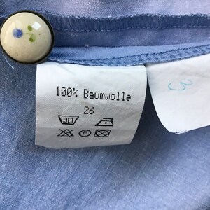 Pu&ograve; includere: Un'etichetta bianca con testo nero e simboli. L'etichetta dice "100% Baumwolle" e ha la taglia 26. Ci sono simboli per il lavaggio, l'asciugatura e la stiratura.