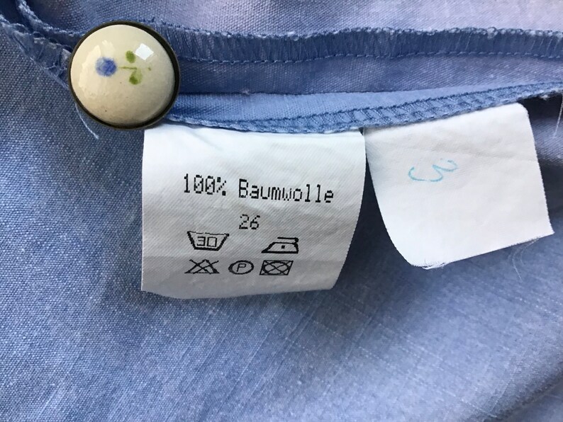 Pu&ograve; includere: Un'etichetta bianca con testo nero e simboli. L'etichetta dice "100% Baumwolle" e ha la taglia 26. Ci sono simboli per il lavaggio, l'asciugatura e la stiratura.