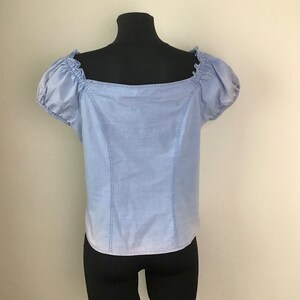 Pu&ograve; includere: Un top in denim blu chiaro con scollo quadrato e maniche a sbuffo corte. Il top &egrave; su un manichino nero.
