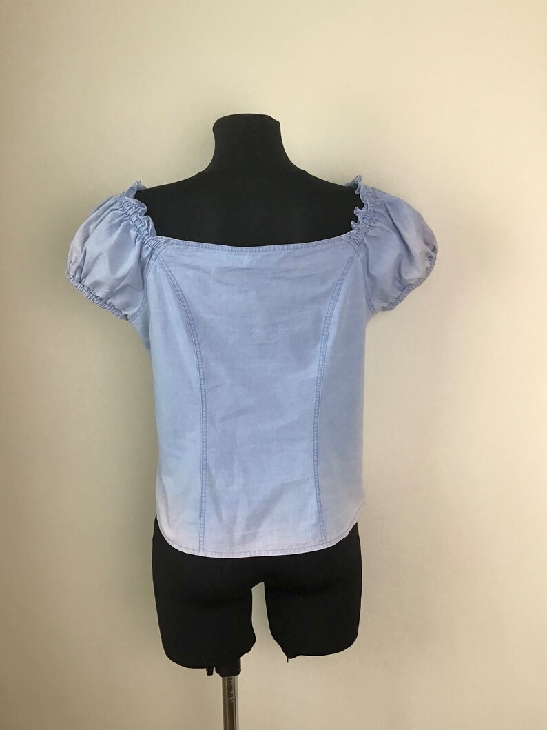 Pu&ograve; includere: Un top in denim blu chiaro con scollo quadrato e maniche a sbuffo corte. Il top &egrave; su un manichino nero.