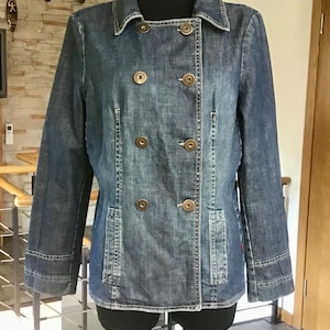 Peut inclure: Veste en jean à double boutonnage. La veste a un col classique, des manches longues et deux poches avant. Elle est fermée par de gros boutons ronds de couleur bronze. La veste est présentée sur un mannequin noir.