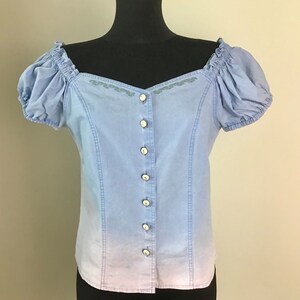 Pu&ograve; includere: Una blusa blu chiaro a spalle scoperte con chiusura a bottoni sul davanti e dettagli ricamati. La blusa presenta un effetto sfumato, che sfuma in una tonalit&agrave; di blu pi&ugrave; chiara nella parte inferiore.