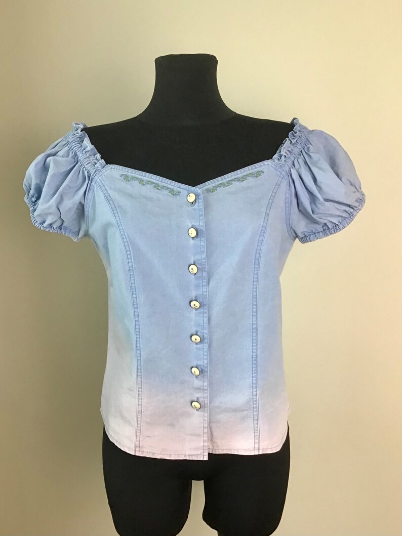 Pu&ograve; includere: Una blusa blu chiaro a spalle scoperte con chiusura a bottoni sul davanti e dettagli ricamati. La blusa presenta un effetto sfumato, che sfuma in una tonalit&agrave; di blu pi&ugrave; chiara nella parte inferiore.