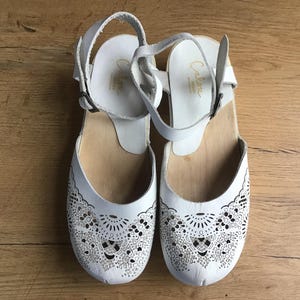 May include: A pair of white leather clogs with decorative cut-out patterns on the toe box. The clogs have wooden soles and ankle straps with buckles. The brand name "Calore" is visible on the insole.
