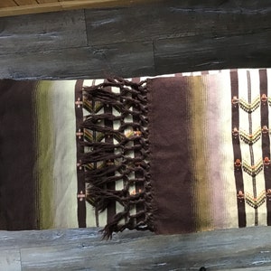 May include: A brown and white striped woven blanket with fringe on the ends. The blanket has a geometric pattern in brown, green, and orange.