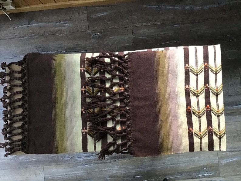 May include: A brown and white striped woven blanket with fringe on the ends. The blanket has a geometric pattern in brown, green, and orange.