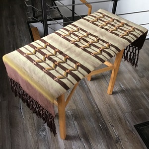 May include: A brown and beige woven table runner with a geometric pattern and fringe on the ends. The runner is laying on a wooden table.