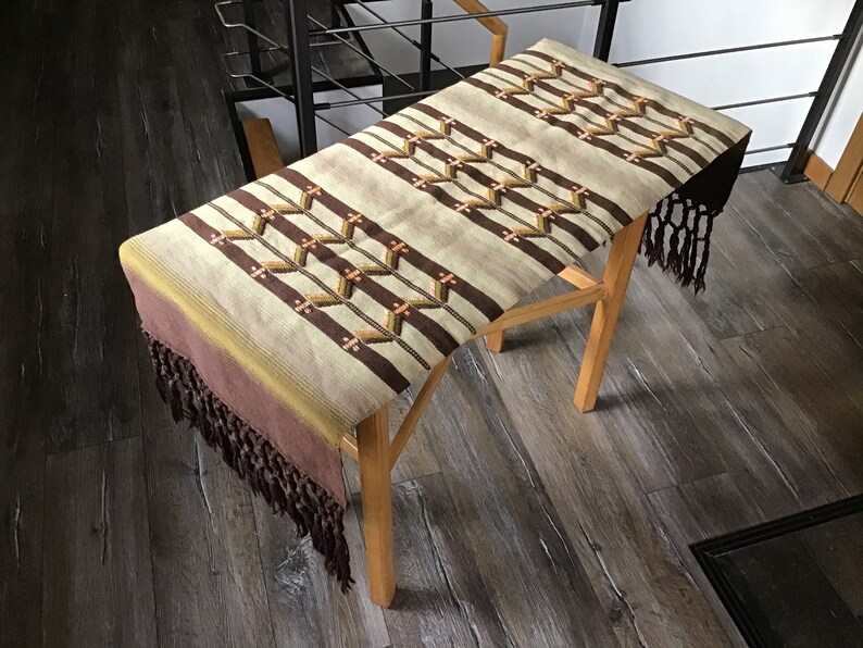 May include: A brown and beige woven table runner with a geometric pattern and fringe on the ends. The runner is laying on a wooden table.