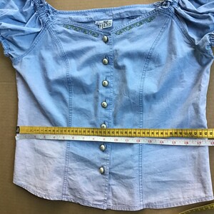 Pu&ograve; includere: Una blusa azzurra abbottonata con maniche corte e dettagli ricamati. La blusa ha un collo a balze e misura 43 cm di larghezza del busto.