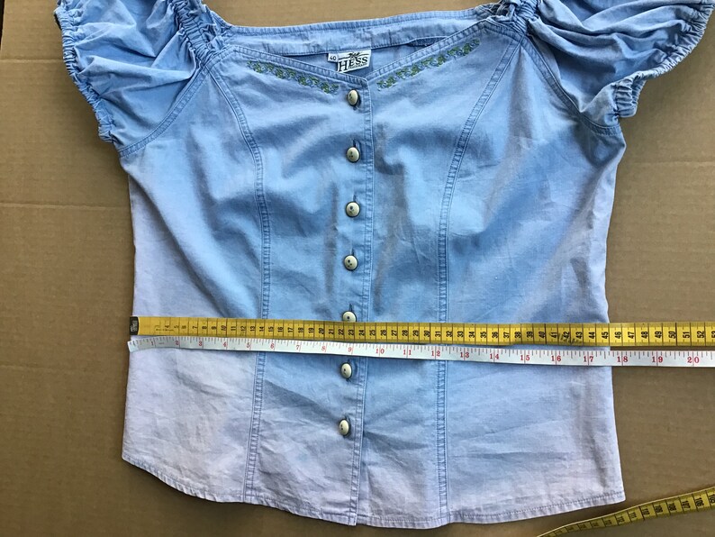 Pu&ograve; includere: Una blusa azzurra abbottonata con maniche corte e dettagli ricamati. La blusa ha un collo a balze e misura 43 cm di larghezza del busto.