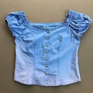 Pu&ograve; includere: Una blusa di colore azzurro chiaro a maniche corte, con chiusura a bottoni sul davanti e un collo ricamato con fiori. La blusa ha una manica leggermente a sbuffo e una vestibilit&agrave; rilassata.