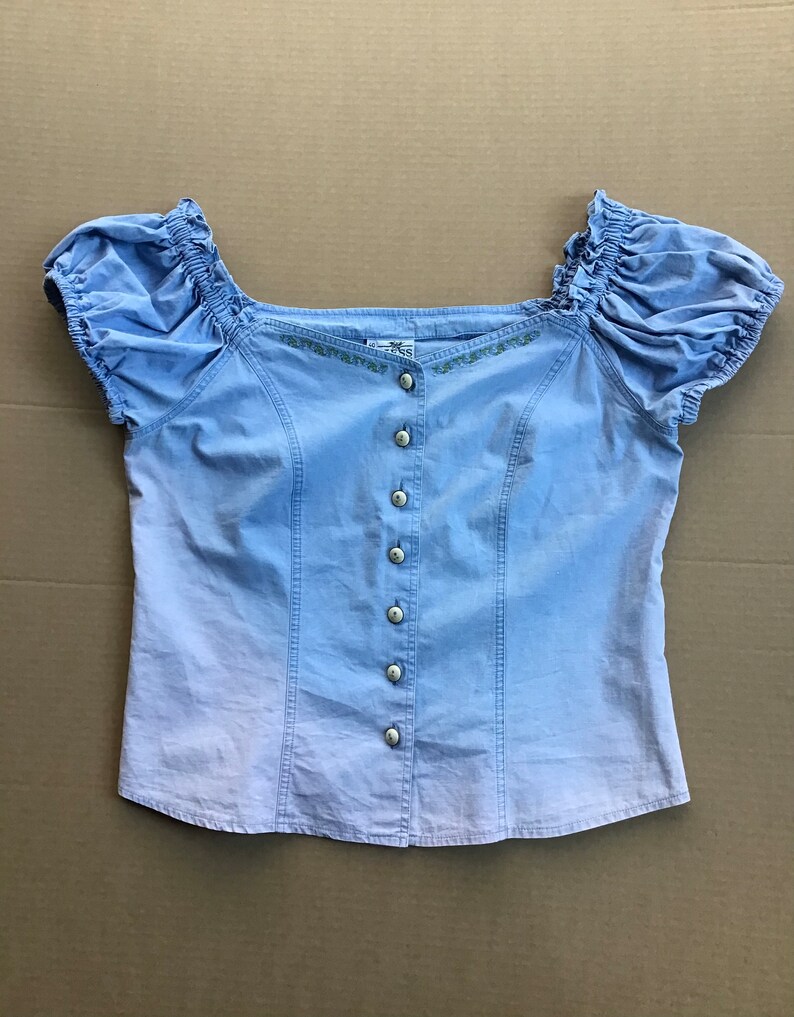 Pu&ograve; includere: Una blusa di colore azzurro chiaro a maniche corte, con chiusura a bottoni sul davanti e un collo ricamato con fiori. La blusa ha una manica leggermente a sbuffo e una vestibilit&agrave; rilassata.