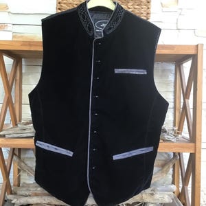 May include: A black velvet vest featuring a high collar with white decorative stitching, a front button closure, and three pockets with grey trim. The garment is displayed on a hanger and includes a label inside the collar with visible text.