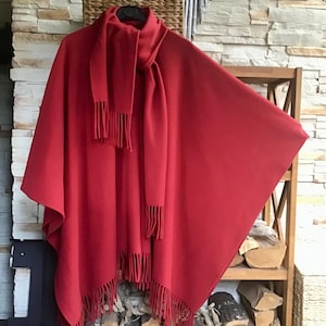 May include: A vibrant red cape and matching scarf with fringed edges. The cape is draped over a hanger, showcasing its flowing design. The scarf is wrapped around the neck of the cape. The ensemble is displayed against a rustic backdrop.