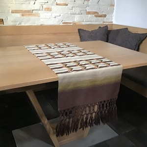 May include: A brown table runner with a geometric pattern and fringe. The runner is on a light wood table with two brown cushions on a bench.