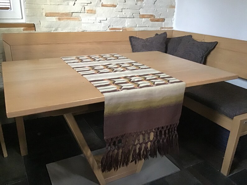 May include: A brown table runner with a geometric pattern and fringe. The runner is on a light wood table with two brown cushions on a bench.