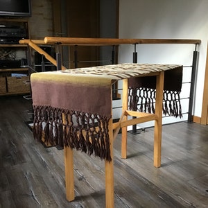 May include: A brown and beige woven table runner with fringe on the ends. The runner is draped over a wooden table with a black metal railing in the background.