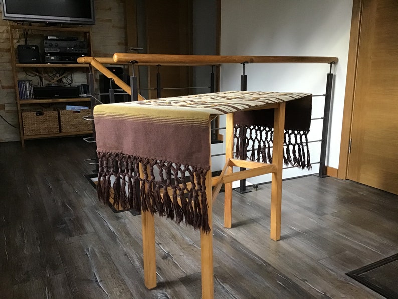 May include: A brown and beige woven table runner with fringe on the ends. The runner is draped over a wooden table with a black metal railing in the background.