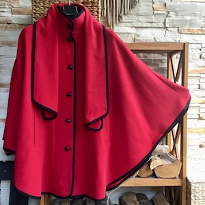 May include: A bright red cape with a high collar and black trim along the edges. The garment features a front button closure with five black buttons and a wide, draped collar design that extends down the front, creating a layered appearance.