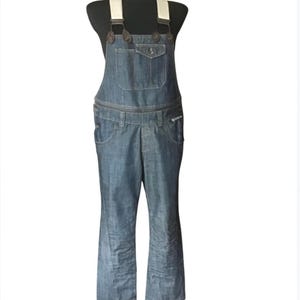 May include: Blue denim dungarees with cream straps and brown buttons. The dungarees feature a bib pocket, side pockets, and a zip closure. The legs are flared at the bottom. The garment is displayed on a black mannequin.