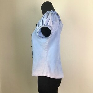 Pu&ograve; includere: Una blusa a maniche corte di colore azzurro chiaro con scollo arricciato e maniche a sbuffo. La blusa presenta un effetto sfumato, passando da un blu pi&ugrave; scuro nella parte superiore a un blu pi&ugrave; chiaro nella parte inferiore.
