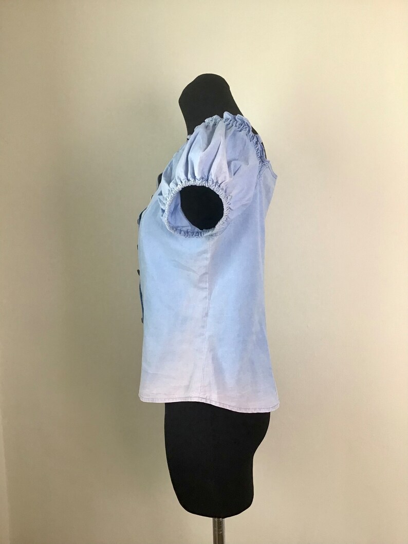 Pu&ograve; includere: Una blusa a maniche corte di colore azzurro chiaro con scollo arricciato e maniche a sbuffo. La blusa presenta un effetto sfumato, passando da un blu pi&ugrave; scuro nella parte superiore a un blu pi&ugrave; chiaro nella parte inferiore.
