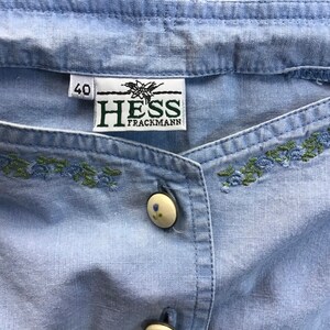 Pu&ograve; includere: Una camicia di cotone blu chiaro con un bottone bianco e ricami verdi e blu. La camicia ha un'etichetta che dice "HESS TRACMANN" e "40".