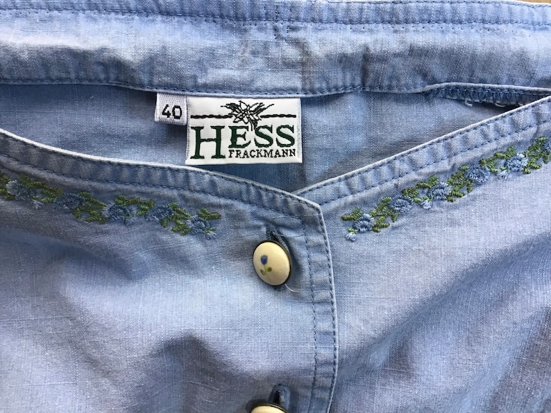 Pu&ograve; includere: Una camicia di cotone blu chiaro con un bottone bianco e ricami verdi e blu. La camicia ha un'etichetta che dice "HESS TRACMANN" e "40".