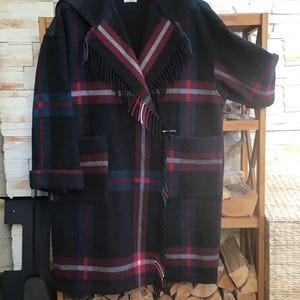 May include: A black hooded coat featuring a plaid pattern in red, blue, and gray stripes. The coat has a wide collar with fringe detailing, long sleeves with folded cuffs, and two large front pockets. A toggle closure secures the front.