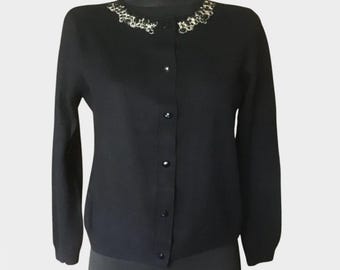 May include: A black long-sleeve cardigan featuring a round neckline adorned with silver-toned metallic embellishments and clear crystals. The garment has a front button-down closure with five matching black buttons and a straight hemline.