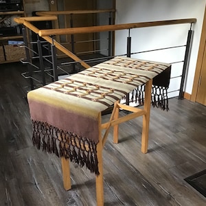 May include: A brown and beige woven table runner with fringe on the ends. The runner is draped over a wooden table stand.