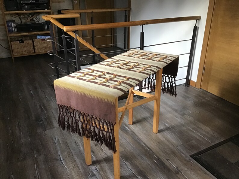 May include: A brown and beige woven table runner with fringe on the ends. The runner is draped over a wooden table stand.
