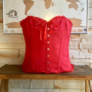 May include: A red satin corset with a lace trim and a bow at the neckline. The corset has a busk closure and is adorned with silver-toned metal eyelets.