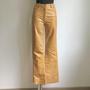 May include: Tan leather pants with a high waist and a straight leg. The pants have a button and zipper closure and pockets. The pants are displayed on a mannequin with a black top.
