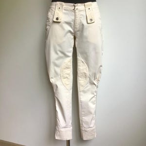Puede incluir: Pantalones de montar de color crema con rodilleras reforzadas y un cierre de botón único en la cintura. Los pantalones tienen un ajuste a medida, con una longitud ligeramente recortada y un sutil puño en el dobladillo. La tela parece ser una mezcla de algodón resistente.