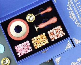 May include: A blue storage box containing a wax seal kit with a gold and wood melting spoon, a wood-handled metal stamp, a round gold-toned melting furnace, and three rectangular containers filled with pink, gold, and copper-colored wax beads.