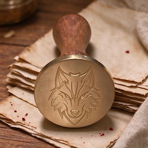 Könnte beinhalten: Ein kreisförmiger Wachssiegelstempel aus Messing mit einer eingravierten Wolfskopf-Gestaltung. Der Stempel hat eine polierte Metallfläche und einen glatten, abgerundeten Holzgriff. Die Wolfsgravur zeigt detaillierte Gesichtszüge und spitze Ohren.