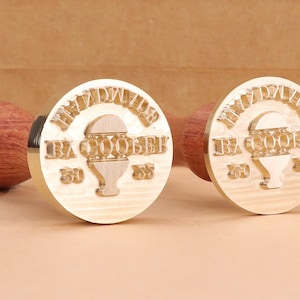 May include: Two wooden stamps with round, light-colored stamp heads and wooden handles. The stamp heads feature the text "HANDMADE" arched across the top, with "BA GOOBEE" and numbers below. The handles are a reddish-brown color.