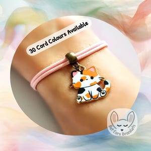 May include: A light pink cord bracelet featuring a metal charm of a calico cat with orange, black, and white enamel details. The charm is attached to the cord with a bronze-toned metal bail. The text 30 Cord Colours Available is visible.