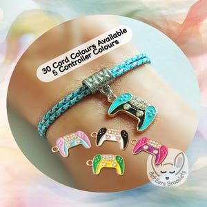 May include: A braided blue and teal cord bracelet featuring a silver-toned metal bead and a dangling video game controller charm with turquoise and black enamel and clear rhinestones. Additional controller charms in pink, white, black, and yellow are shown.