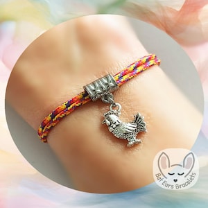 May include: A colorful bracelet with a rooster charm. The bracelet features a woven cord in red, yellow, and purple. The charm is a detailed silver-colored rooster. The bracelet is worn on a wrist.